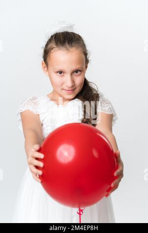 Front view of playful girl holding out red balloon with both hands and looking at camera. Studio shot Caucasian girl 10 years old on white background Stock Photo