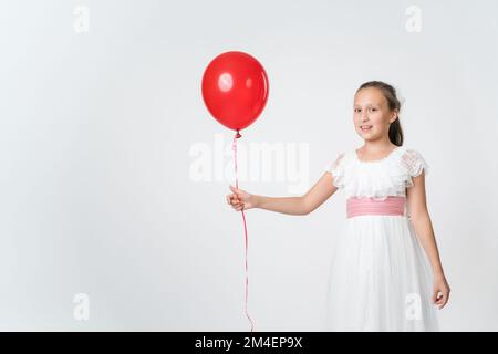 Smiling girl dressed long white dress holding one red balloons in hand, smiling looking at camera. Studio shot of Caucasian girl of 10 years old Stock Photo