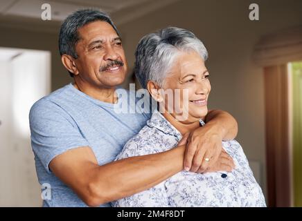 Everything we do, we do together. a cheerful mature couple holding each other while looking outside through a window at home during the day. Stock Photo