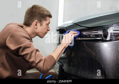 Car wash and cleaning at professional auto service station. Close up view of caucasian young male worker cleaning car headlights with blue microfiber cloth using high pressure water. Stock Photo