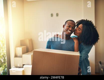 Thank you for doing the heavy lifting. Portrait of a cheerful young man carrying a cardboard box while his girlfriend gives him a kiss on the cheek. Stock Photo