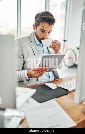 Linked to the latest business updates. a handsome young businessman drinking coffee while working on a digital tablet in an office. Stock Photo