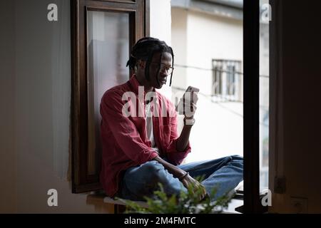 Young African guy sitting on windowsill freelancing with smartphone from home Stock Photo
