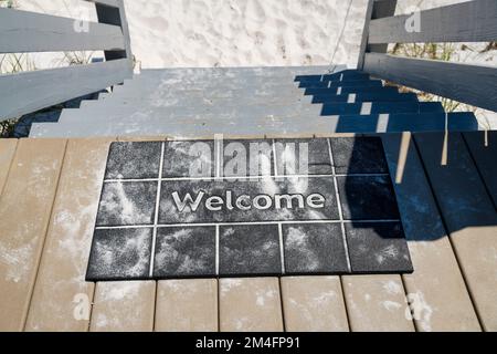 Destin, Florida- Black welcome mat with white sand on top of a wooden stairs. High angle view of a mat on a wood planks flooring near the steps of the Stock Photo