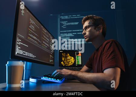 Side view portrait of modern IT developer using computer at home office at night, working on software, coding new application, sitting in front of big Stock Photo