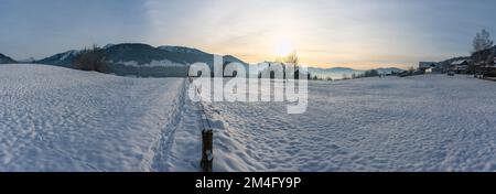 Panorama mit verschneiter Landschaft, Schnee auf Wiese, Dorf, einzelner Baum und die Berge des Bregenzerwald im Hintergrund. Abendsonne über Hittisau Stock Photo