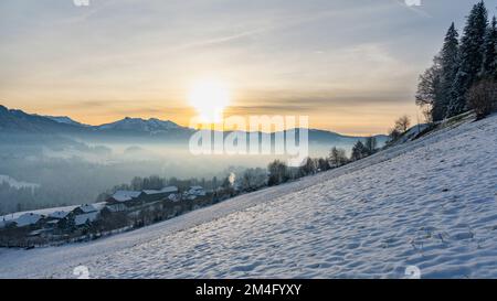 Panorama mit verschneiter Landschaft, Schnee auf Wiese, Dorf, einzelner Baum und die Berge des Bregenzerwald im Hintergrund. Abendsonne über Hittisau Stock Photo