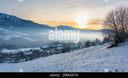 Panorama mit verschneiter Landschaft, Schnee auf Wiese, Dorf, einzelner Baum und die Berge des Bregenzerwald im Hintergrund. Abendsonne über Hittisau Stock Photo