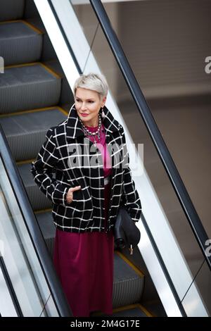 Fashionable stylish mature woman with gray hair standing on escalator, wearing bright image of plaid jacket and long burgundy dress Stock Photo