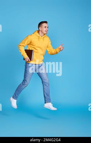 jumping very excited man with book in yellow sweatshirt and jeans isolated on blue background. Full length Stock Photo