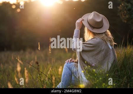Happy woman with hat and poncho relaxing outdoors during sunset. Mental wellbeing and resting in nature Stock Photo
