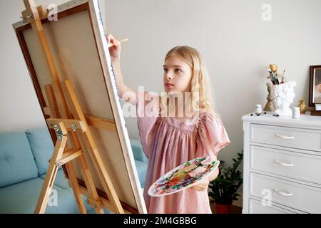 Beautiful blonde teenage girl artist in pink dress painting picture on canvas on easel, holding palette with paints at bright home interior Stock Photo