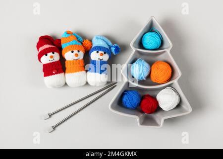 Three knitted snowmen in blue, orange and red hats with balls of thread and knitting needles on a gray background. Winter knitting creativity, the con Stock Photo