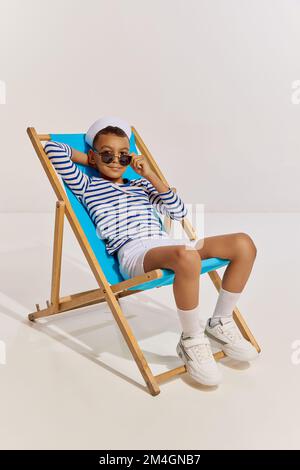 Portrait of boy, child in striped vest and sunglasses sitting on chair, posing over grey background. Cruise travelling Stock Photo