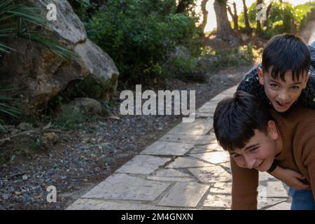 Funny boy climbing on back of cheerful teen brother while spending time on paved path at sundown in park Stock Photo