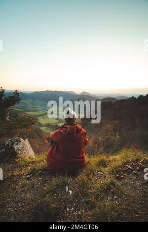 Enthusiastic traveller sitting on the edge of a rock in a red jacket during sunset. Sulovske skaly, Slovakia, Eastern Europe. Stock Photo