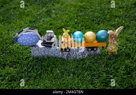 models of toy gray excavator and yellow dump truck stand on granite stone in grass along with bright colorful Easter eggs. Easter spring holiday conce Stock Photo