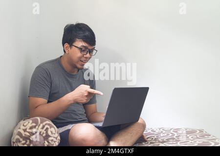 Portrait Asian eye glasses man is pointing and looking at laptop on a bedroom for work from home concept with natural light Stock Photo