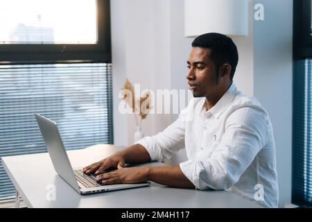 Side view of confident young African-American male entrepreneur working typing on business project on laptop sitting at desk in modern office room. Stock Photo