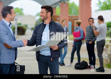 two mature men shaking hands on campus students in background Stock Photo