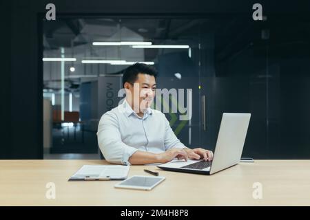 Portrait of a young Asian teacher sitting in the office at the table. He works with a laptop, teaches online, remotely. Recruits, conducts classes via video call, smiles at students. Stock Photo