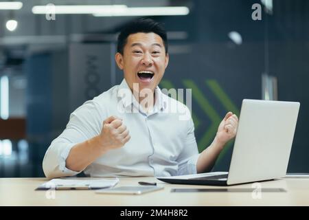 A young Asian businessman plays online games at work in the office with a laptop, places bets. He is happy to win, shouts with happiness, looks at the camera. Stock Photo