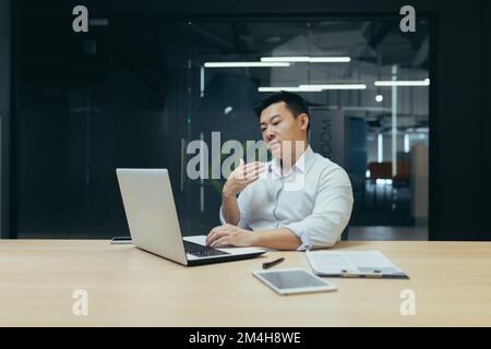 A young Asian man is sitting in the office at the table, working on a laptop. He waves his hand in his face. He feels bad, feels hot, needs fresh air, medical help. Stock Photo