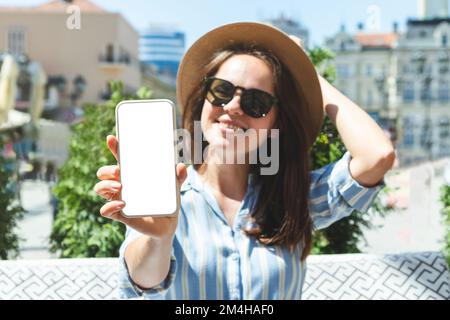 Woman in sun hat holding smartphone in hand and showing blank screen at camera. Stock Photo