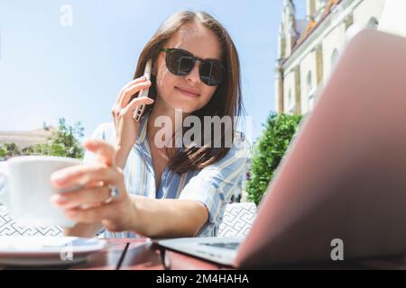 Young brunette businesswoman talking mobile phone and using laptop while sitting in outdoor cafe and drinking coffee. Stock Photo
