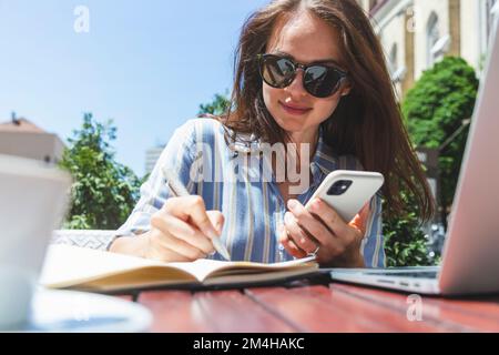 Businesswoman in sunglasses writing notes in her work notebook and using her mobile phone while sitting in outdoor cafe in summer. Stock Photo