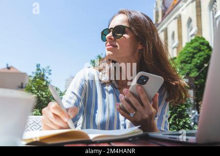 Businesswoman in sunglasses writing notes in her work notebook and using her mobile phone while sitting in outdoor cafe in summer. Stock Photo