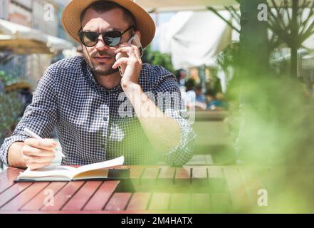 Serious man in a sun hat and sunglasses talking mobile phone and writing notes in a notebook while sitting outdoors cafe. Stock Photo