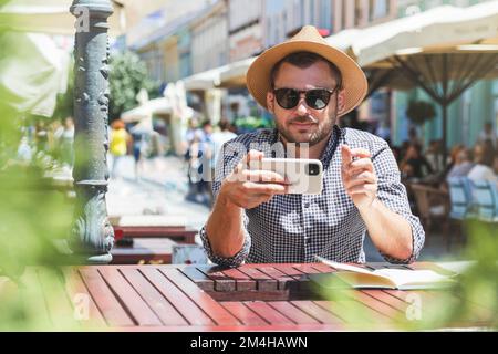 Man in sun hat using smartphone while sitting in outdoor cafe in the city. Stock Photo