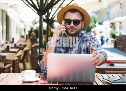 Casual man using laptop and calling on the mobile phone while sitting in outdoor cafe. Stock Photo