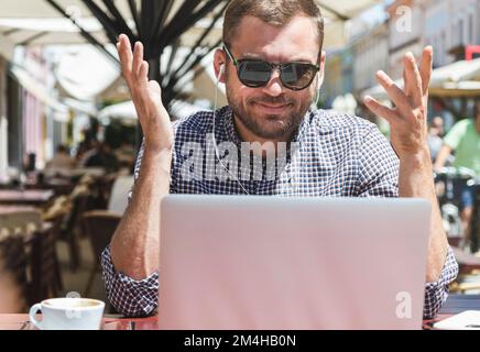Handsome adult man in sunglasses talking video call using his headphones and laptop while sitting in an outdoors cafe. Stock Photo