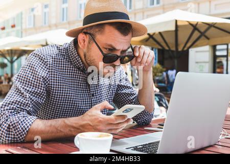 Man in sun hat staring at the laptop screen and holding smartphone in hand. Stock Photo