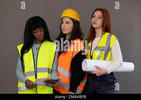 Three models of different ethnicities posing, dressed as construction workers. Reflective vests, hard hats and rolled up project. Stock Photo