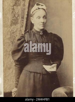 Antique photograph of victorian lady stood near curtains and a chair holding a letter. wearing a bonnet and a broach on her high collar.Fashion Stock Photo