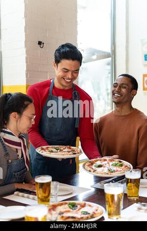 Waiter serving delicious pizza to a group of friends sitting in a casual cafe. The waiter holding two pizzas in his hand putting it on the table. Drin Stock Photo