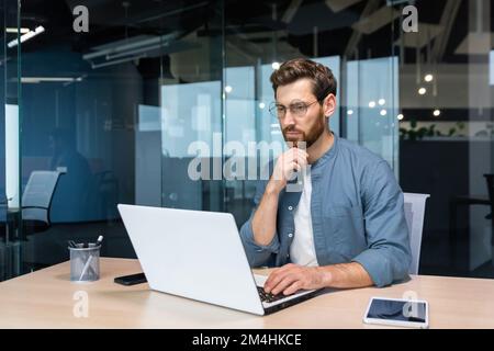 .Serious pensive businessman in shirt thinking about decision sitting at table in modern office, man with beard is using laptop at work. Stock Photo
