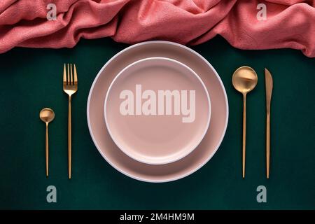 Festive place setting with pink napkin. Empty plates and gold cutlery on dark green background. Top view. Dinner table in luxury restaurant. Card or m Stock Photo