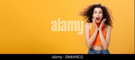 Waist-up shot of charmed enthusiastic attractive female with curly hairstyle in cropped top holding palms on cheeks and smiling astonished and Stock Photo