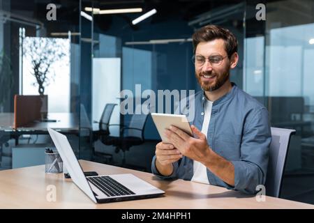 Successful businessman in a shirt uses a tablet computer, a man sits at a desk smiling and happy, a programmer tests a new application. Stock Photo