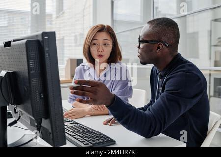 African American programmer pointing at computer monitor and discussing computer program with his colleague Stock Photo