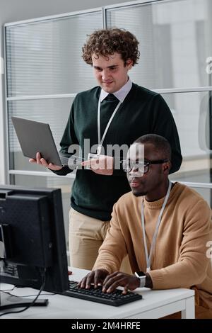 African American developer typing codes on keyboard with his colleague standing in background and checking codes on his laptop Stock Photo