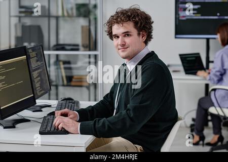 Portrait of young IT specialist looking at camera while working at his workplace and typing codes on computer Stock Photo