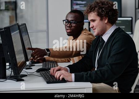 Programmer pointing at computer monitor with codes and talking to his colleague while they working with computer program in team Stock Photo