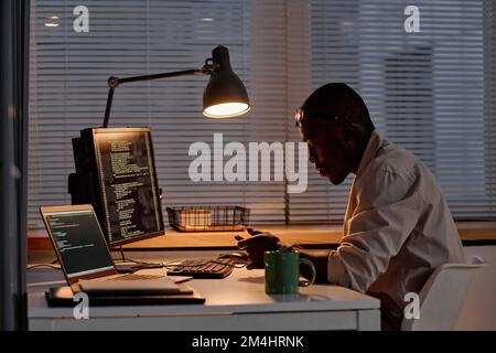 African American IT specialist using his smartphone while working with codes on computer at his workplace till late at night Stock Photo