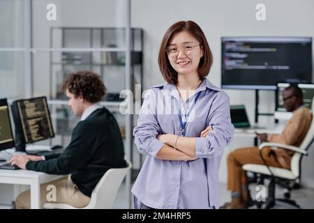 Portrait of Asian young woman smiling at camera standing with her arms crossed in IT office with her colleagues in background Stock Photo