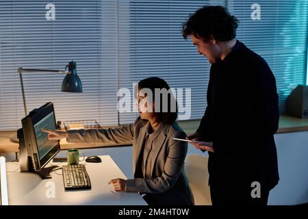 Young woman pointing at computer monitor with codes and discussing their security with her colleague in office Stock Photo